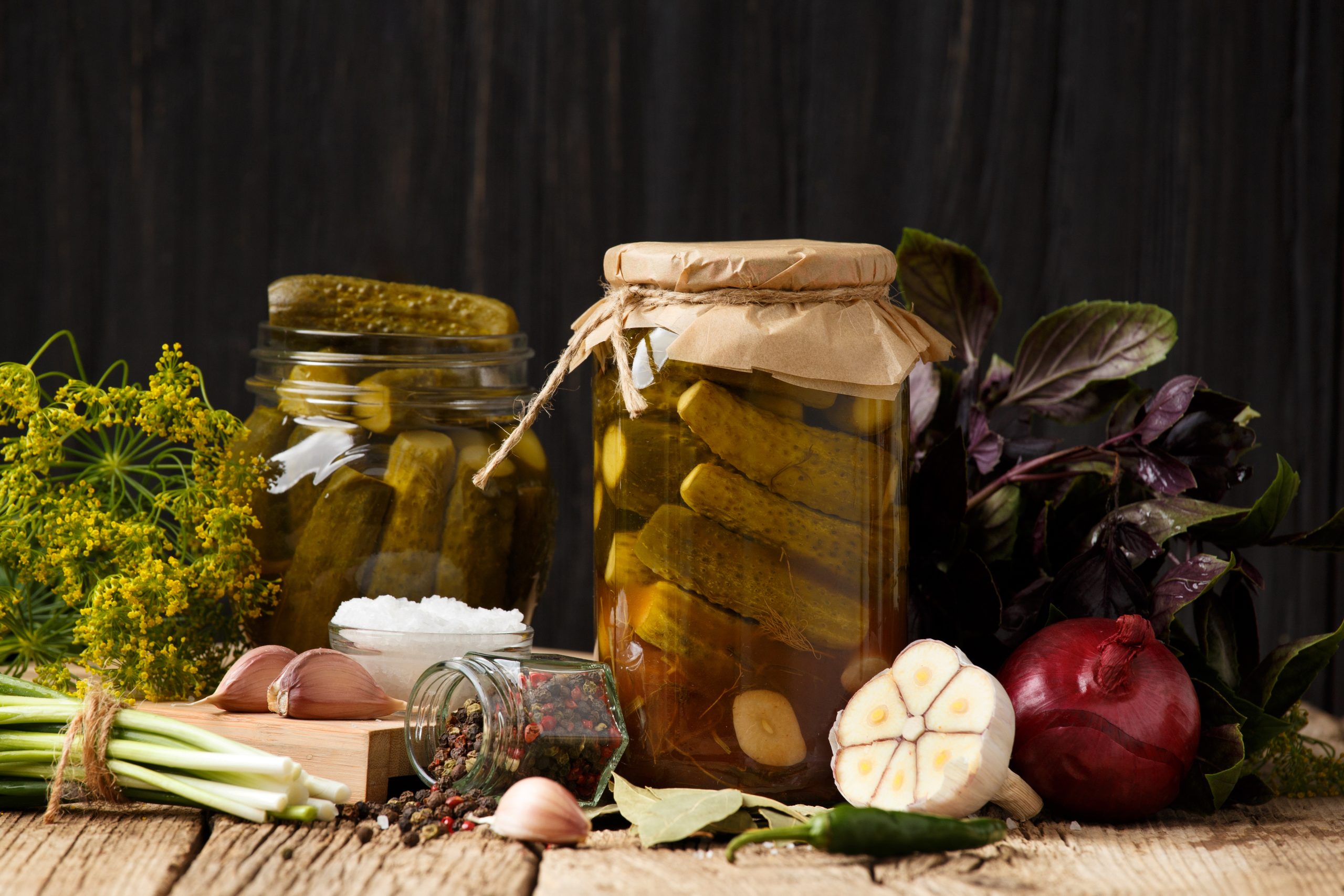 Pickled cucumbers in closed and open jars, onion, garlic, dill, parsley, basil on a dark wooden background. Homemade canned gherkins , spices and herbs for marinade.